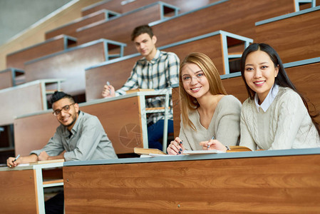 多种族群体的人坐在现代大学教室的讲堂的不同桌子上背景图片