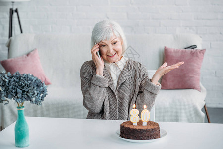 快乐的年长女子在家中与生日蛋糕一起坐在桌边聊天时用智能手图片