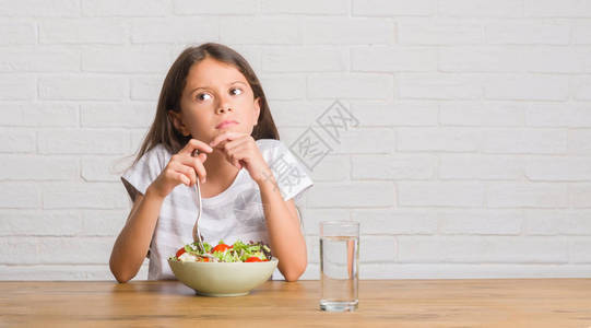 坐在桌子上吃健康沙拉的年轻的西班牙小孩面对着严肃的面孔思考问题非图片