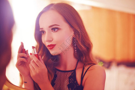 一个美丽的年轻女子正在做自己的改造用红光在镜子前用口红做晚妆的cerly头发的女孩关图片
