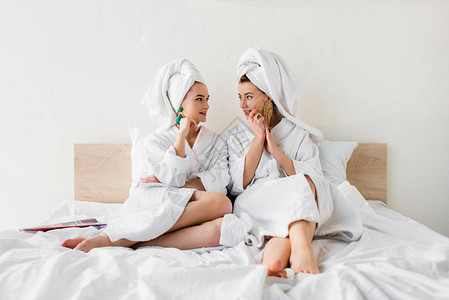 戴耳环浴袍和头部毛巾的时髦女孩坐在床上互背景图片