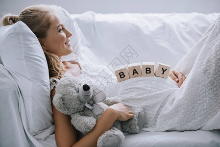 身穿白色睡衣带着泰迪熊和木块的微笑孕妇的侧视图图片