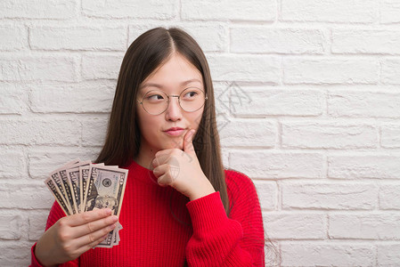 年轻女人在砖墙上拿着一美元严肃的面子思考问题图片