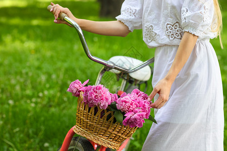 美丽的金发女人穿着漂亮的裙子在公园里骑着一篮美丽的牡丹花图片