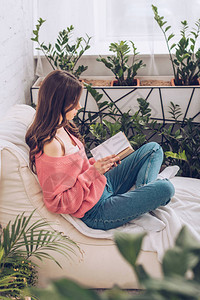 年轻女在家里的绿色植物附近盘腿坐着看书图片