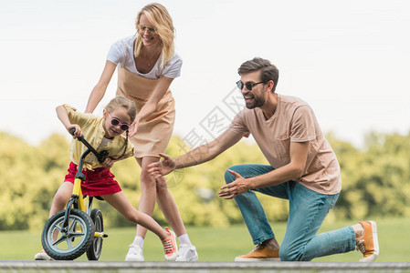 快乐的父母看着可爱的小女儿在公园骑自行图片