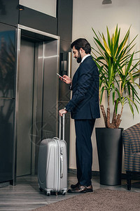 商人在旅馆等候电梯时携带使用智能电话的行李图片