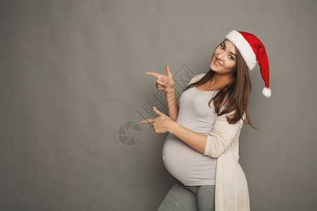 圣诞老人帽子的年轻美丽的孕妇指向旁边画象图片