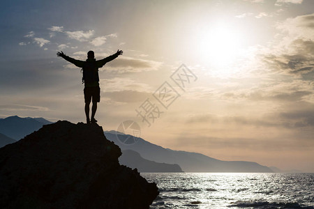 男人张开双臂庆祝或在美丽的鼓舞人心的日出与山脉和大海男子徒步旅行或举起双手在克里特岛的岩石顶部享受鼓背景图片