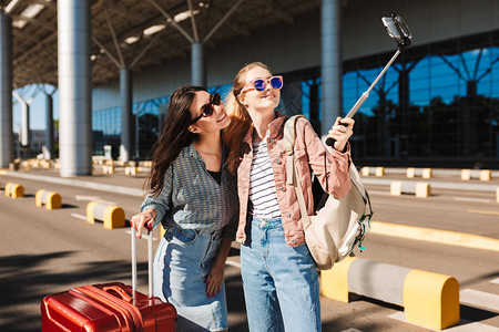 两个戴着墨镜的漂亮女孩在机场附近的户外肩上带着手提箱和背包图片