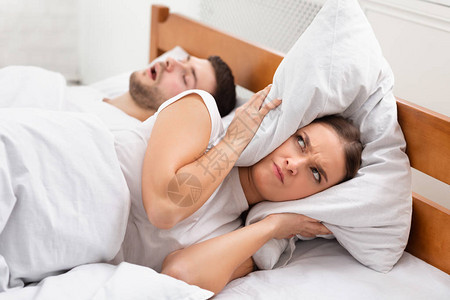 打鼾愤怒的女朋友用枕头在家里睡觉的男朋友附近捂着耳朵图片