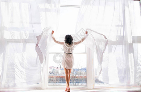 年轻女子的照片站在敞开的窗帘之间的窗户上图片