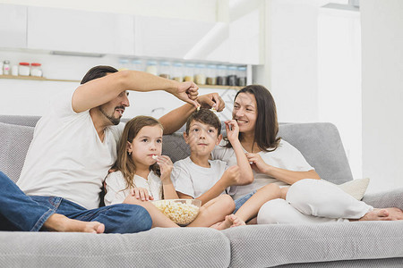 美丽的年轻父母及其子女正在看电视吃爆米花和在家中坐在沙发上微笑图片