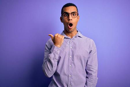 英俊的非洲裔美国人穿着条纹衬衫和紫色背景的眼镜惊讶地用手指着一边图片