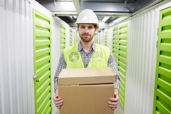 戴工作头盔的留着胡子的年轻人拿着纸板箱在储藏室工作时看图片