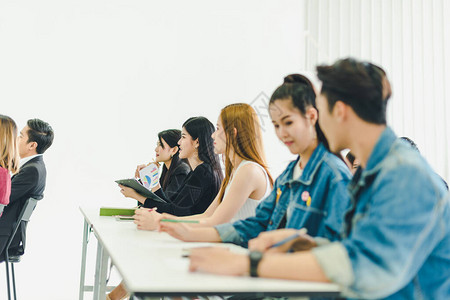 亚洲人参加研讨会并在培训室聆听演讲者的演讲有些人会记笔有人举手询问解说员并在演讲背景图片
