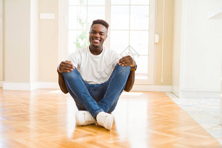 英俊的非洲男人坐在家里的地板上面带快乐和凉爽的笑图片