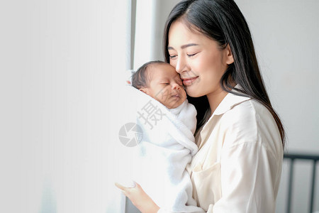 白衬衫亚洲母亲在卧室里用白色窗帘在玻璃窗前亲吻她刚出生的婴儿图片