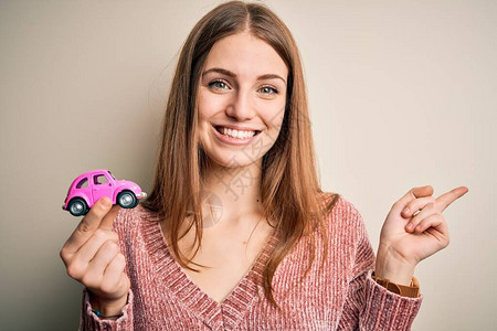 年轻漂亮的红发美女拿着小粉红色车在孤立的白种背景上以手和指着图片