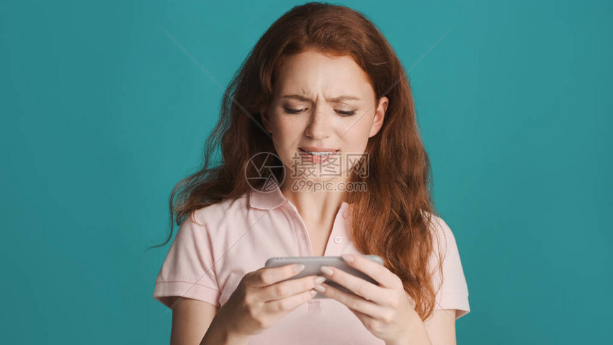 有吸引力的神经紧张的红发女孩在多彩背景上玩智能手机游戏图片