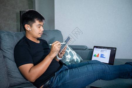 亚洲商人使用无线耳机笔记本和平板在家工作他为在家中举行的网上会议准备图表和信息校图片