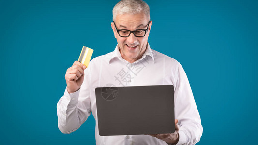 轻松付款兴奋的戴眼镜的老人拿着信用卡和笔记本电脑在蓝色工作室图片