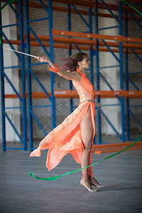 穿着橙色衣服的年轻身穿橙色裙子在仓库中手握体操丝带跳舞的图片