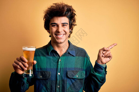 英俊的年轻帅哥喝着啤酒杯站在孤立的黄色背景上以手和指向另图片