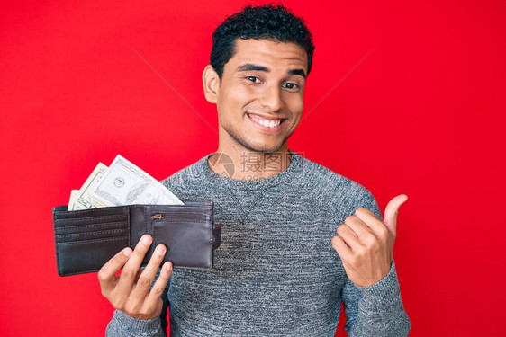 年轻英俊的西班牙小帅哥拿着钱包用美元指着大拇指向侧面笑着图片
