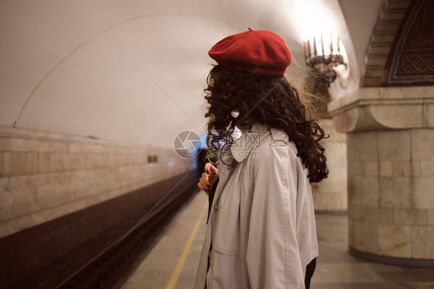 穿着风衣和红色贝雷帽在地铁站等车的时尚黑发女孩的侧视图图片