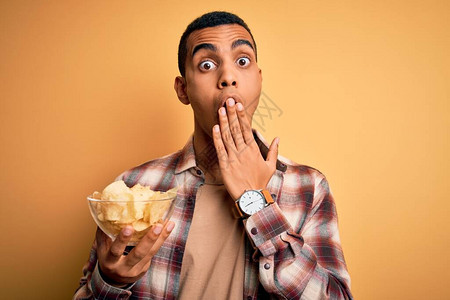 年轻英俊的非洲裔美国人拿着一碗薯片盖在黄色背景上图片