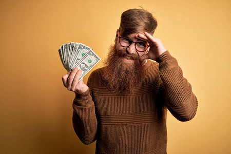 留着胡子的爱尔兰红发男子拿着一堆1美元钞票在黄色背景上背景图片