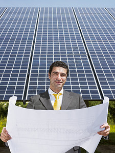 成人中意大利男工程师在太阳能发电站阅读蓝图并微笑的肖像图片