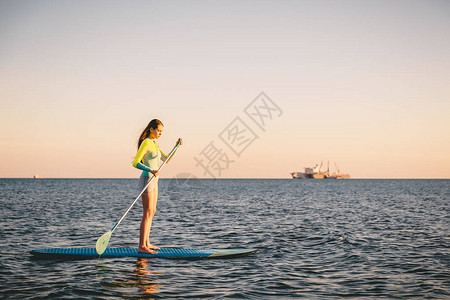 女孩在黄昏时站起船桨登机在平坦温暖宁静的海面上图片