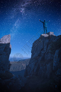 站在岩石悬崖边缘的人手举起来与夜景对峙在星般的天空中图片