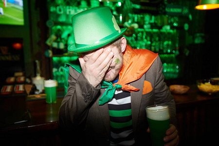 戴着绿帽子的肮脏的黑衣男子用杯啤酒在另一面图片