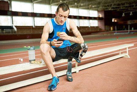 年轻截肢运动员脱离在长椅上吃三明治和使用智能手机练习时脱机的完整图片
