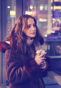 一个穿着带兜帽的保暖夹克的年轻女孩用纸杯和热咖啡暖手城市生活模图片