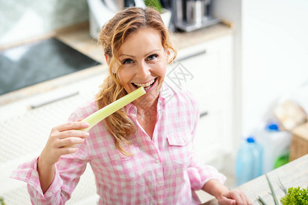 微笑的女人在厨房里拿着并品尝新鲜的绿色芹菜图片
