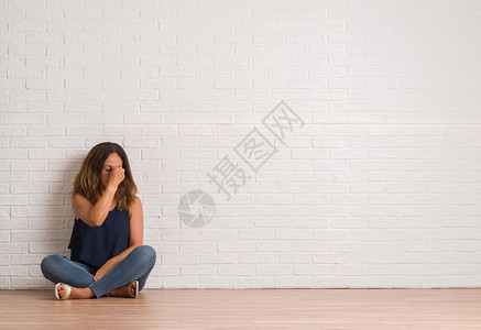 坐在白砖墙上地板上的中年女精神失常者感到疲惫疲倦鼻涕和眼神疲劳背景图片