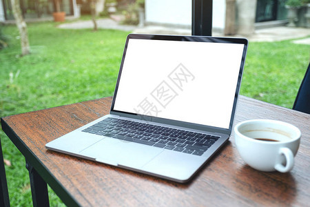 用白色空白桌面屏幕和绿色自然背景的木桌边咖啡杯制成笔记本图片