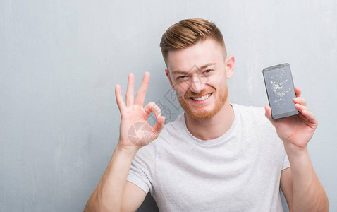 灰色格朗基墙上的年轻红发男子拿着破碎的智能手机用手指做ok标志图片