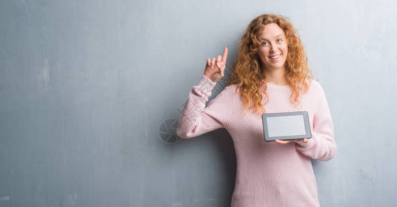 红发女青年在灰墙上使用平板电脑惊讶于一个想法或问题用快乐的面孔指图片