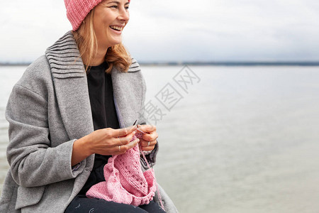 一位身着时髦外套的年轻已婚妇女用针头对着大海编织一顶粉红色的帽子图片
