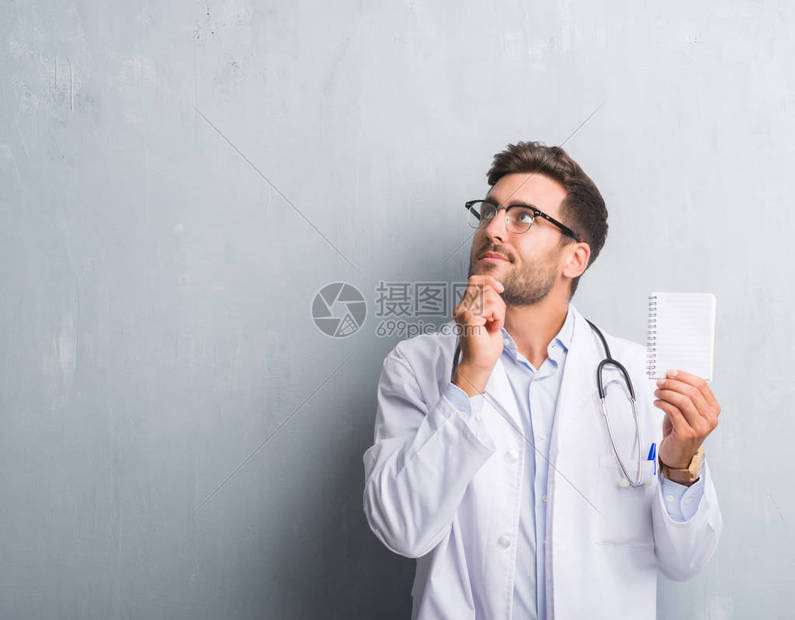 年轻帅气的医生男人在灰墙上拿着空白笔记本严肃地面对着问题图片