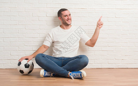 坐在白砖墙边的年轻caucasian男子拿着足球非常快乐地用手和图片