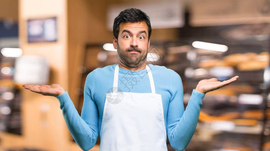 穿着围裙的男人在举手和肩膀时怀疑和困惑的表情在面包店里图片