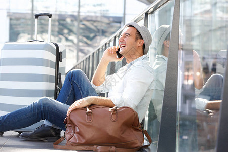 坐在地板上带着手提袋和在车站用移动电话交谈的快乐旅行男图片