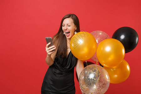 快乐的女人穿着黑色小礼服尖叫着拿气球用手机庆祝孤立在红色上情人节快乐新年生日模拟背景图片