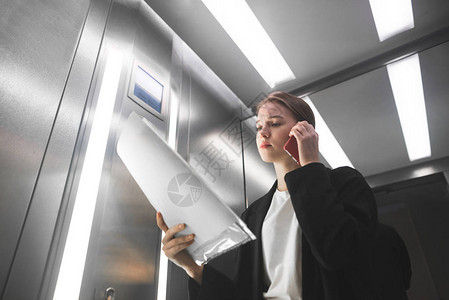 忧心忡的女上班族正在打电话和检查电梯里的文件年轻的女商人在电梯里听智能手机和图片